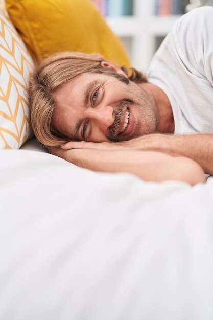 Junger blonder Mann lächelt selbstbewusst und liegt im Schlafzimmer auf dem Bett