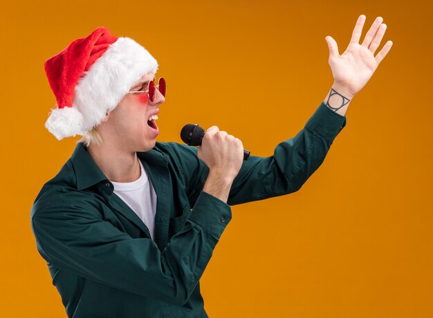 Junger blonder Mann mit Weihnachtsmütze und Brille mit Mikrofon, der auf die Seite schaut und die Hand in der Luft singt, isoliert auf orangefarbenem Hintergrund