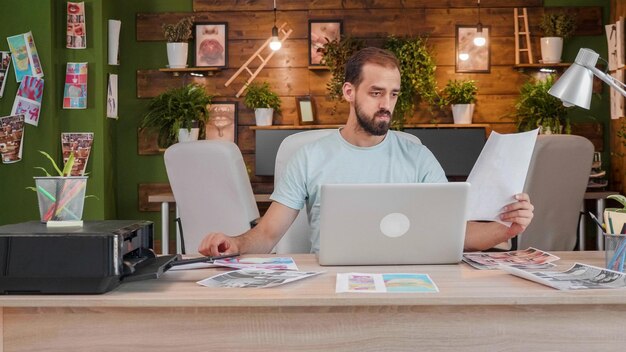 Junger Designer, der ein Blatt betrachtet, das es in der Hand hält, während er am Tisch vor einem Laptop sitzt. Kaukasischer Kreativer