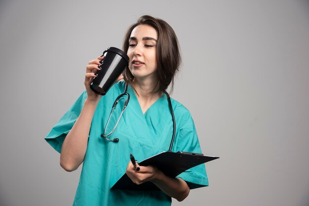 Junger Doktor, der Kaffee auf grauem Hintergrund trinkt. Hochwertiges Foto