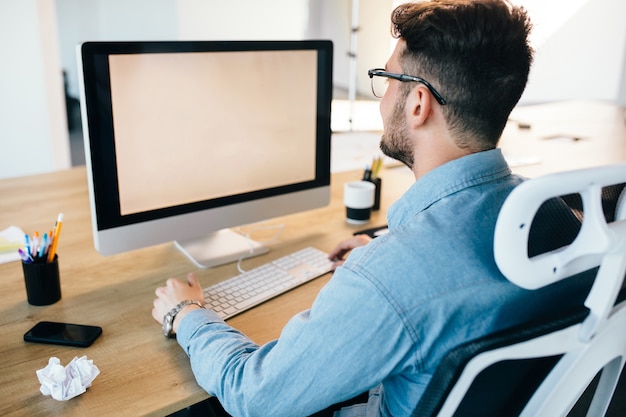 Junger dunkelhaariger Mann arbeitet mit einem Computer an seinem Schreibtisch im Büro. Er trägt ein blaues Hemd und sieht beschäftigt aus. Blick von hinten.