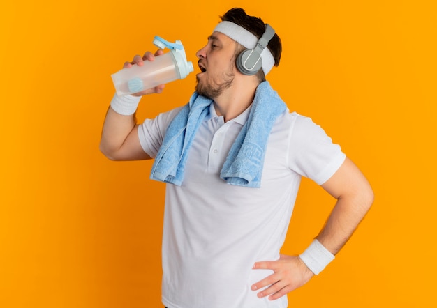 Junger eignungsmann im weißen hemd mit dem stirnband und dem handtuch um hals, der flasche trinkwasser nach dem training steht über orange hintergrund hält