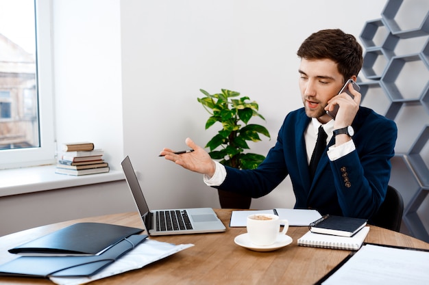 Junger erfolgreicher geschäftsmann, der am telefon, bürohintergrund spricht.