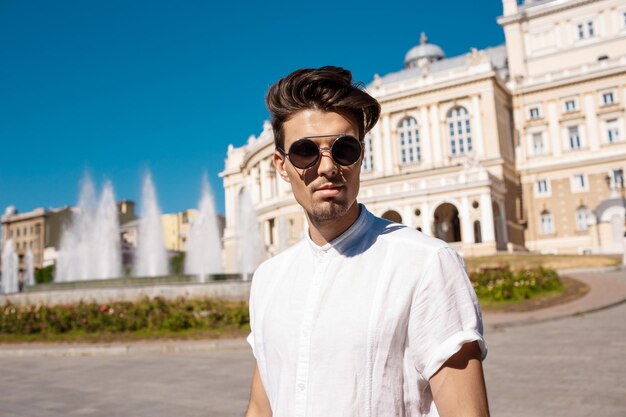 Junger erfolgreicher Geschäftsmann mit Sonnenbrille, der lächelnd durch die sonnige Stadt geht