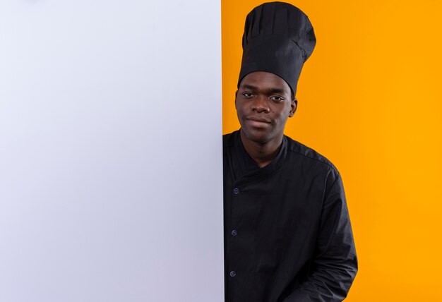 Junger erfreuter afroamerikanischer Koch in der Kochuniform steht hinter weißer Wand lokalisiert auf orange Raum mit Kopienraum