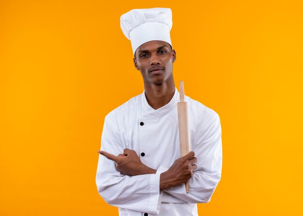 Junger erfreuter afroamerikanischer Koch in der Kochuniform verschränkt die Arme und hält Nudelholz isoliert auf orange Wand