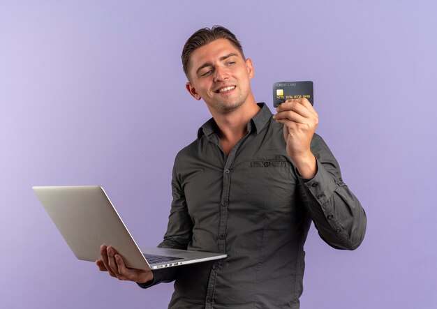 junger erfreuter blonder hübscher Mann hält Laptop und schaut auf Kreditkarte