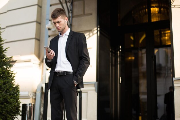 Junger ernsthafter Geschäftsmann im schwarzen Anzug und im weißen Hemd mit drahtlosen Kopfhörern, die nachdenklich das Handy im Freien verwenden