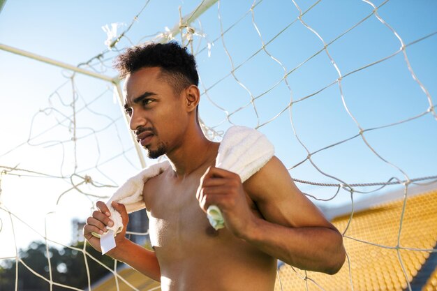 Junger ernsthafter, hemdloser afroamerikanischer Sportler mit Handtuch auf den Schultern nach dem Fußballspiel im Stadtstadion