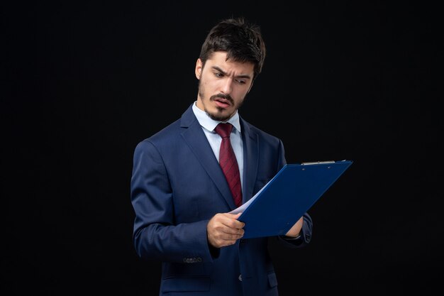 Junger ernsthafter männlicher Büroangestellter im Anzug, der Dokumente hält und Statistiken an isolierter dunkler Wand überprüft