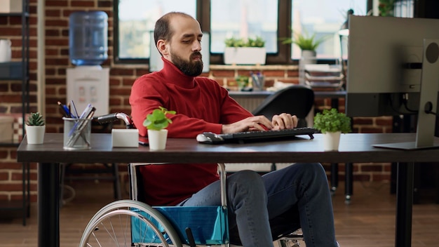 Junger Erwachsener mit Behinderung, der an Finanzanalysen arbeitet und Computer verwendet, um Strategien für die Geschäftsentwicklung zu erstellen. Mitarbeiter des Unternehmens im Rollstuhlplanungswirtschaftswachstum im Büro.