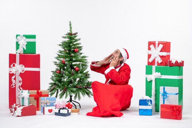 Junger Erwachsener verkleidet als Weihnachtsmann mit Geschenken und geschmücktem Weihnachtsbaum, der im Boden sitzt und mit jemandem in Überraschung spricht