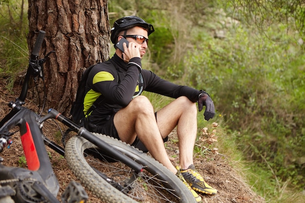 Junger europäischer Radfahrer in der Sportbekleidung, die in den Wäldern ruht, die auf Handy sprechen
