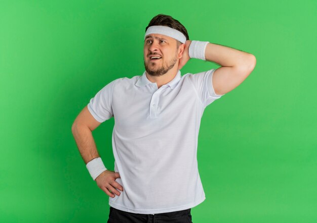 Junger Fitness-Mann im weißen Hemd mit dem Stirnband, das beiseite schaut, verwirrt mit Hand auf Kopf, der über grünem Hintergrund steht