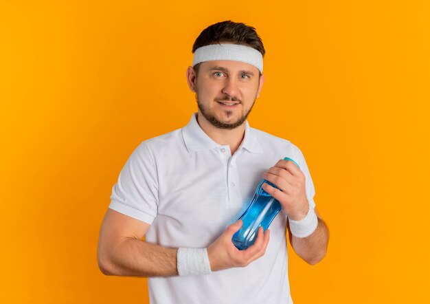 Junger Fitness-Mann im weißen Hemd mit dem Stirnband, das Flasche Wasser hält, das Kamera mit Lächeln auf Gesicht steht, das über orange Hintergrund steht