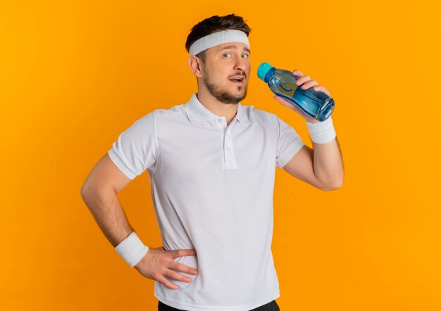 Junger Fitness-Mann im weißen Hemd mit dem Stirnband, das Flasche Wasser hält, das Kamera mit Lächeln auf Gesicht steht, das über orange Hintergrund steht