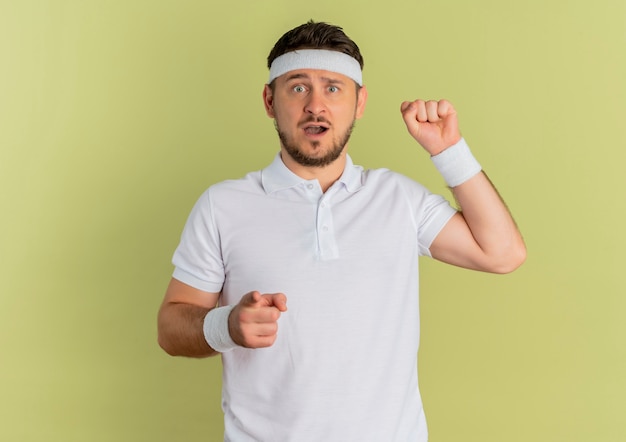 Junger Fitness-Mann im weißen Hemd mit dem Stirnband, das mit dem Finger auf die vordere anhebende Faust zeigt, die überrascht steht über Olivenwand steht