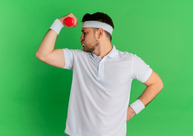 Junger Fitness-Mann im weißen Hemd mit dem Stirnband, das mit der Hantel arbeitet, die über grünem Wand angespannt und zuversichtlich steht