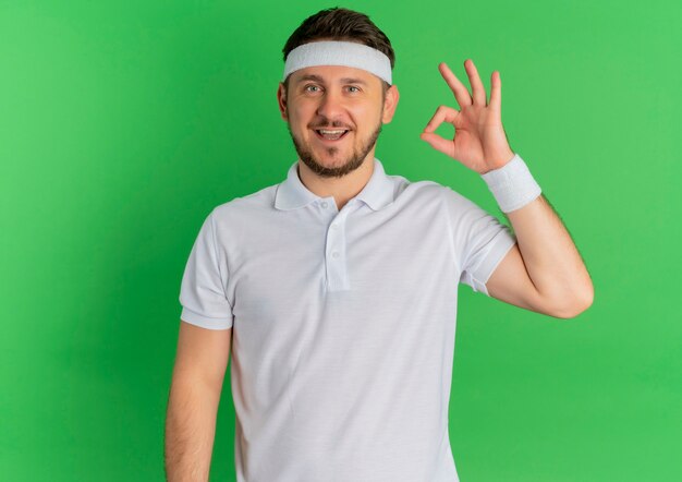 Junger Fitness-Mann im weißen Hemd mit dem Stirnband, das nach vorne lächelnd fröhlich tut, das ok singt, das über grüner Wand steht