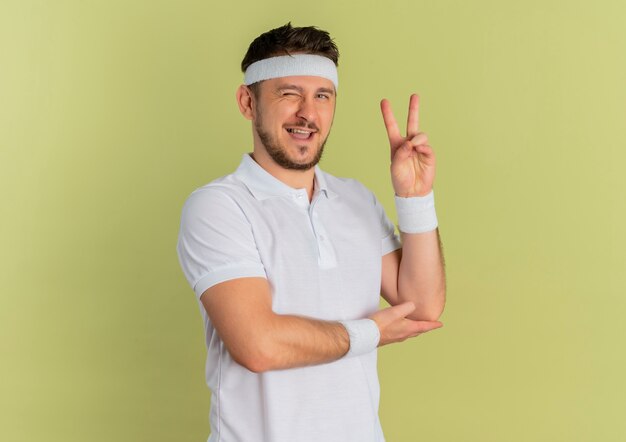 Junger Fitness-Mann im weißen Hemd mit dem Stirnband, das nach vorne lächelt und zwinkert und Siegeszeichen zeigt, das über Olivenwand steht