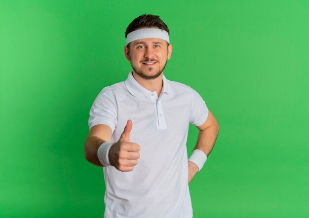 Junger Fitness-Mann im weißen Hemd mit dem Stirnband, das nach vorne mit Lächeln auf Gesicht schaut, das Daumen oben steht über grüner Wand zeigt