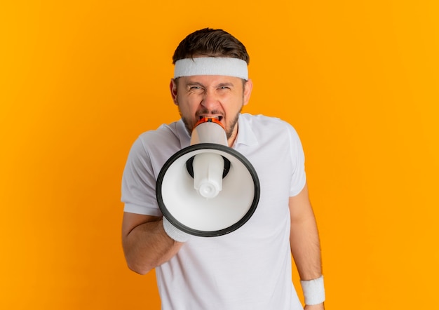 Junger Fitness-Mann im weißen Hemd mit dem Stirnband, das zum Megaphon mit aggressivem Ausdruck schreit, der über orange Wand steht