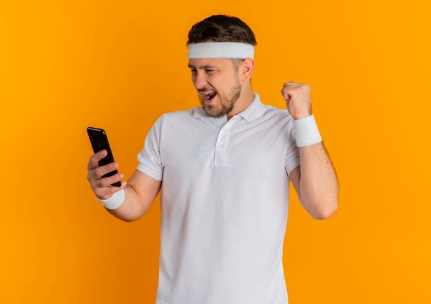 Junger Fitness-Mann im weißen Hemd mit dem Stirnband, der das Smartphone vergoldet, fummelt die Faust glücklich und aufgeregt, über orange Hintergrund stehend