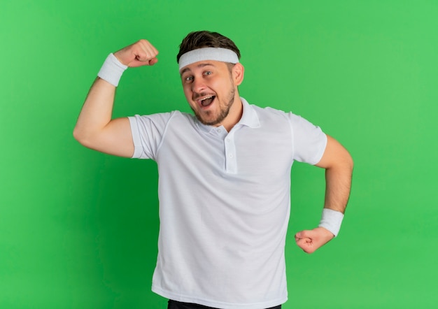 Junger Fitness-Mann im weißen Hemd mit Stirnband, das Faust anhebt, die Bizeps glücklich und aufgeregt zeigt, über grüner Wand stehend