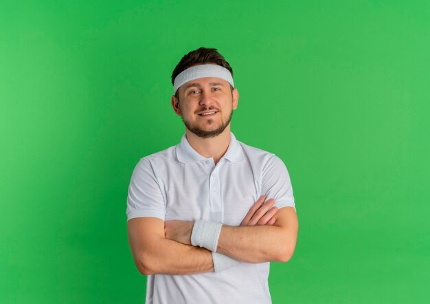 Junger Fitness-Mann im weißen Hemd mit Stirnband mit gekreuzten Händen auf der Brust, die nach vorne mit sicherem Ausdruck über grüner Wand steht