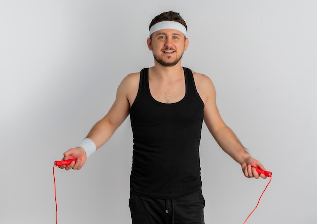 Junger Fitness-Mann mit dem Stirnband, der unter Verwendung des Springseils mit glücklichem Gesicht über weißem Hintergrund springt