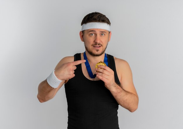 Junger Fitness-Mann mit Stirnband und Goldmedaille um seinen Hals, der mit dem Finger darauf zeigt und zuversichtlich steht, über weißem Hintergrund zu stehen