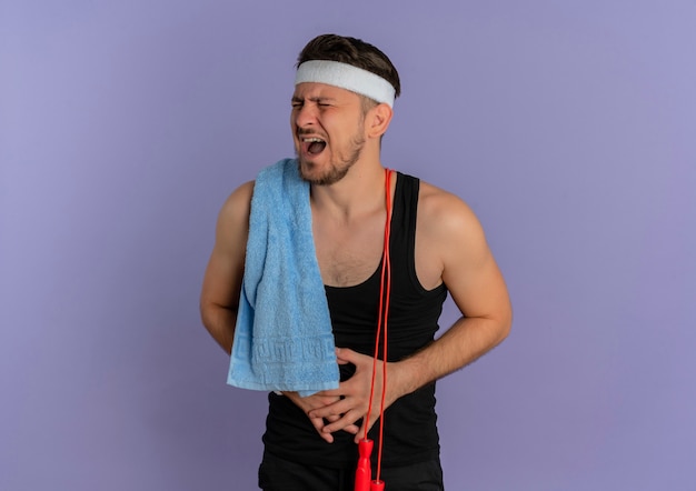 Junger fitness-mann mit stirnband und handtuch auf der schulter, die unwohl suchen, seinen bauch berührend, der unter schmerzen steht, die über lila wand stehen