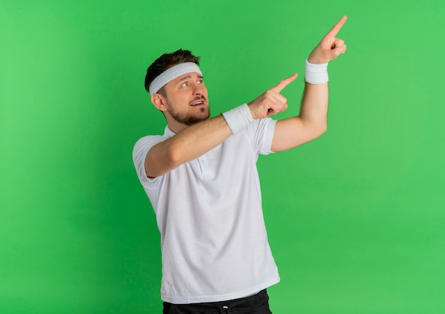 Junger Fitnessmann im weißen Hemd mit dem Stirnband, das beiseite schaut und mit Zeigefingern auf die Seite zeigt, die über grünem Hintergrund steht