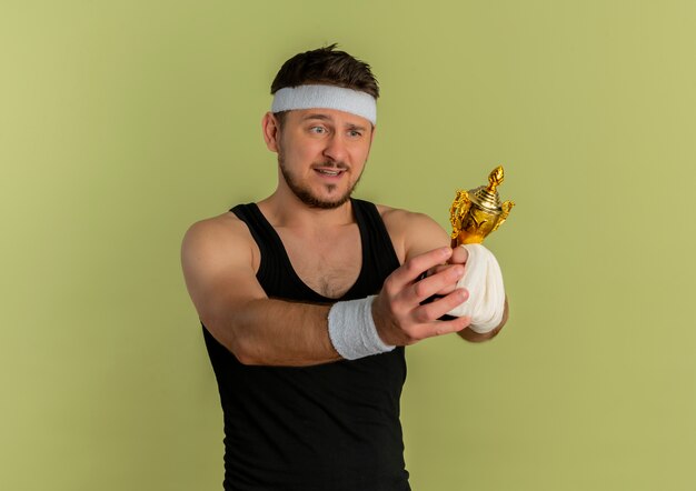 Junger Fitnessmann mit dem Stirnband, der seine Trophäe hält, die es aufgeregt und glücklich betrachtet, über Olivenhintergrund stehend