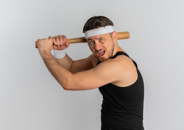 Junger Fitnessmann mit Stirnband, das einen Baseballschläger emotional und aufgeregt schwingt, der über weißem Hintergrund steht