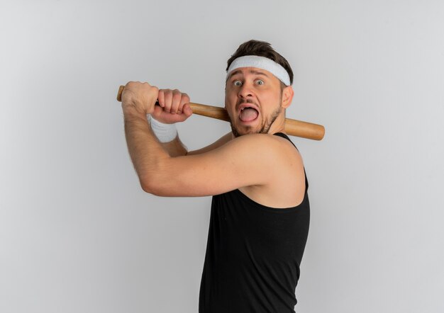 Junger Fitnessmann mit Stirnband und Goldmedaille, die einen Baseballschläger schwingend erschreckten, der über weißem Hintergrund steht