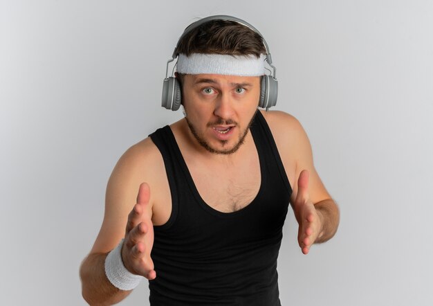 Junger Fitnessmann mit Stirnband und Kopfteilen, die Kamera mit sicherem Ausdruck betrachten, der bereit ist, über weißem Hintergrund zu stehen