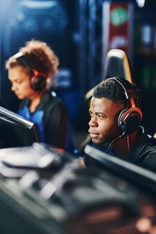 Junger fokussierter afrikaner, cybersport-spieler mit kopfhörern, der am esport-turnier teilnimmt, online-videospiele spielt, während er im internet-café sitzt, vertikaler schuss