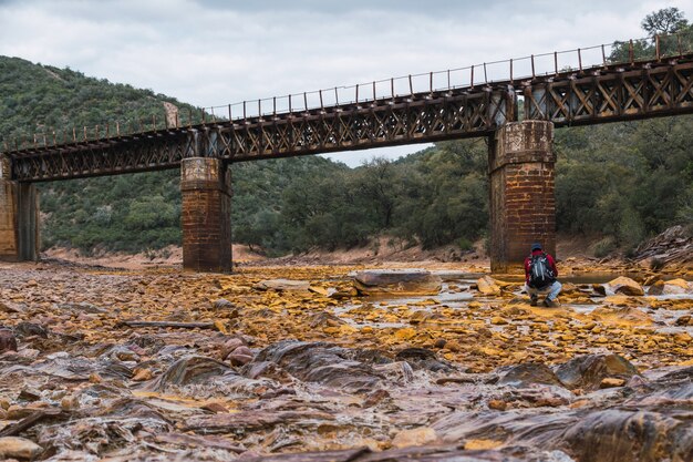 Junger Fotograf mit Rucksack und Mütze beim Fotografieren einer alten Eisenbrücke über den Rio Tinto