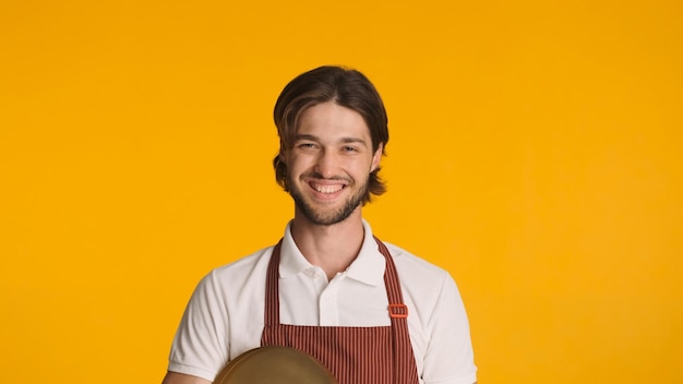 Junger freundlicher Kellner, der die Kamera anschaut und aufrichtig vor einem farbenfrohen Hintergrund lächelt Attraktiver bärtiger Mann in Schürze, der bei der Arbeit glücklich aussieht