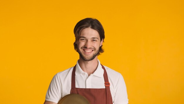 Junger freundlicher Kellner, der die Kamera anschaut und aufrichtig vor einem farbenfrohen Hintergrund lächelt Attraktiver bärtiger Mann in Schürze, der bei der Arbeit glücklich aussieht