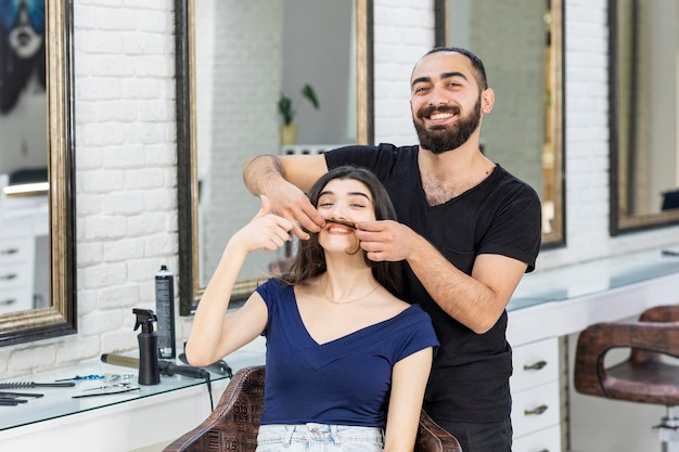 Junger Friseur, der das Haar der Frau als Schnurrbart hält und beide lachen Foto in hoher Qualität