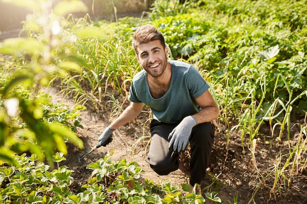 Junger fröhlicher attraktiver bärtiger männlicher Gärtner im blauen T-Shirt und in der schwarzen Sporthose lächelnd, im Garten arbeitend, Sprossen mit Schaufel pflanzend.