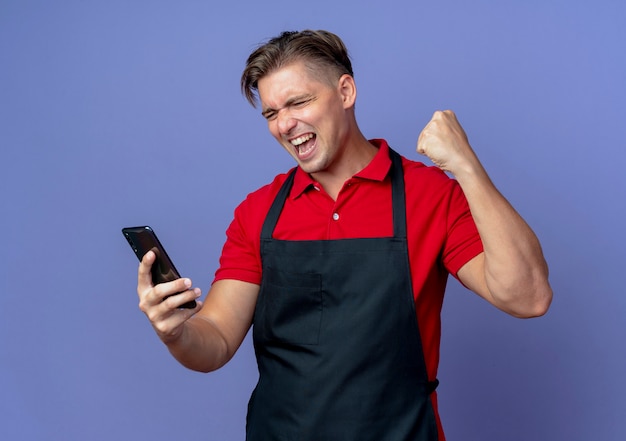 junger fröhlicher blonder männlicher Friseur in Uniform hebt Faust, die Telefon ansieht