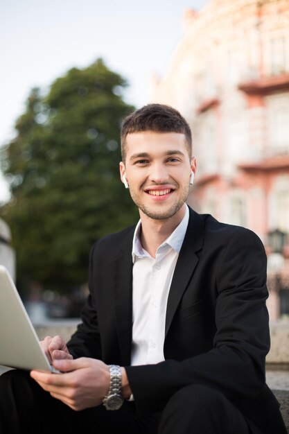 Junger fröhlicher Mann im klassischen schwarzen Anzug und weißem Hemd mit drahtlosen Kopfhörern, der glücklich in die Kamera mit Laptop in den Händen schaut, während er Zeit im Freien verbringt