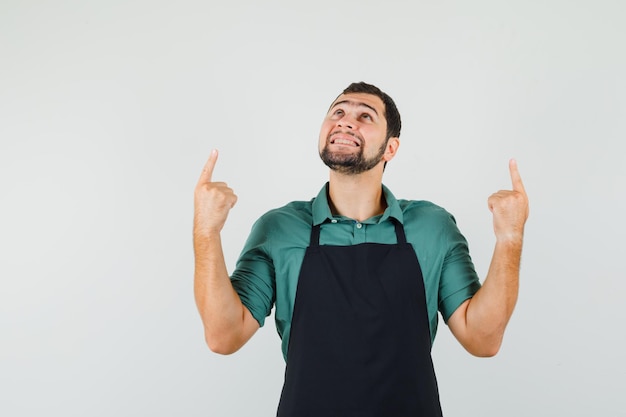 Junger Gärtner, der in T-Shirt, Schürze nach oben zeigt und fröhlich aussieht, Vorderansicht.