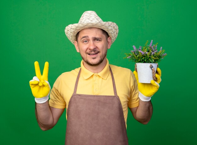 Junger Gärtner, der Overall und Hut in Arbeitshandschuhen trägt, die Topfpflanze stehen über grüner Wand halten
