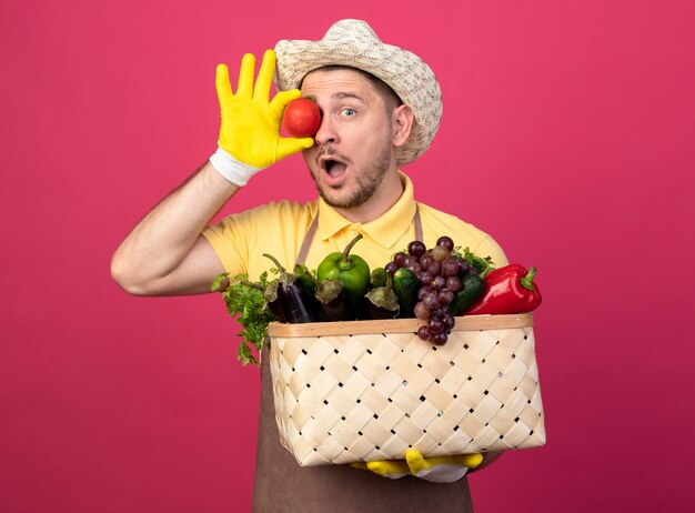 Junger Gärtner, der Overall und Hut in den Arbeitshandschuhen trägt, hält Kiste voll des Gemüses und der frischen Tomate über seinem Auge, das überrascht steht, das über rosa Wand steht