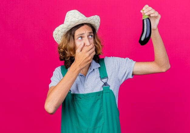 Junger Gärtnermann, der Overall und Hut hält, der frische Aubergine hält