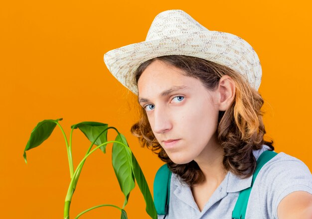 Junger Gärtnermann, der Overall und Hut hält Pflanze tut Selfie, das Kamera schaut, das zuversichtlich steht über orange Hintergrund